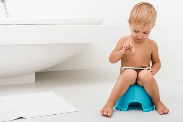 Cute toddler boy sitting on blue potty and using smartphone near bathtub — Stock Photo