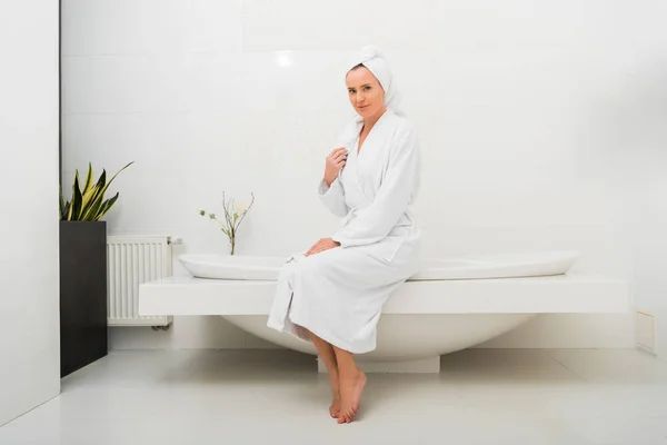 Jolie femme en peignoir blanc assis dans la salle de bain moderne — Photo de stock