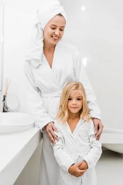 Alegre madre en toalla mirando linda hija en cuarto de baño - foto de stock