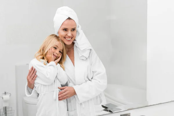 Cheerful mother and cute daughter in in bathrobes looking at mirror in bathroom — Stock Photo
