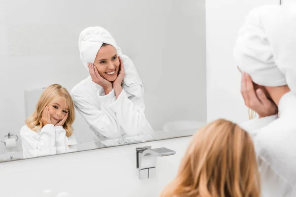 Selective focus of cheerful mother and cute daughter in bathrobes touching faces in bathroom — Stock Photo