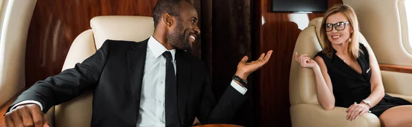 Panoramaaufnahme eines glücklichen afrikanisch-amerikanischen Geschäftsmannes, der im Privatjet in der Nähe einer Geschäftsfrau gestikuliert — Stockfoto