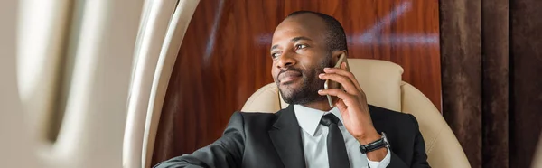 Panoramic shot of handsome african american businessman talking on smartphone in private jet — Stock Photo