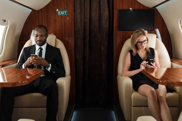 Guapo hombre de negocios afroamericano y atractiva mujer de negocios utilizando teléfonos inteligentes en jet privado - foto de stock