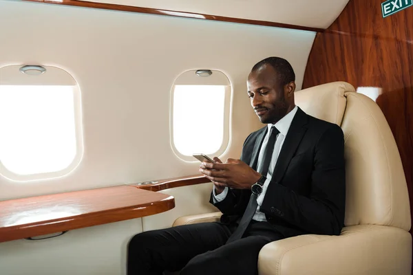 Guapo afroamericano hombre de negocios utilizando teléfono inteligente en jet privado - foto de stock