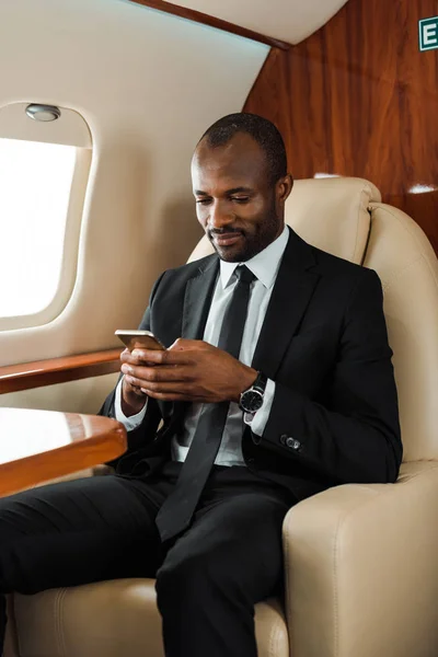 Guapo afroamericano hombre de negocios en traje usando teléfono inteligente en jet privado - foto de stock