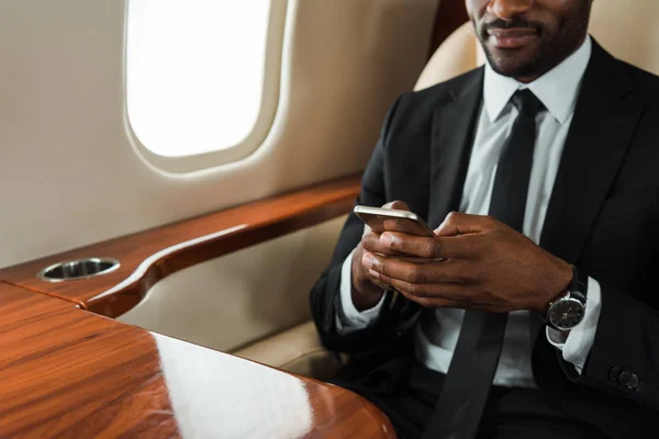 Vista recortada del hombre de negocios afroamericano en traje usando teléfono inteligente en jet privado - foto de stock