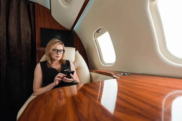 Foyer sélectif de la femme dans les lunettes tenant smartphone en jet privé — Photo de stock