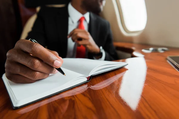 Vista ritagliata dell'uomo africano americano che tiene la penna vicino al notebook in jet privato — Foto stock