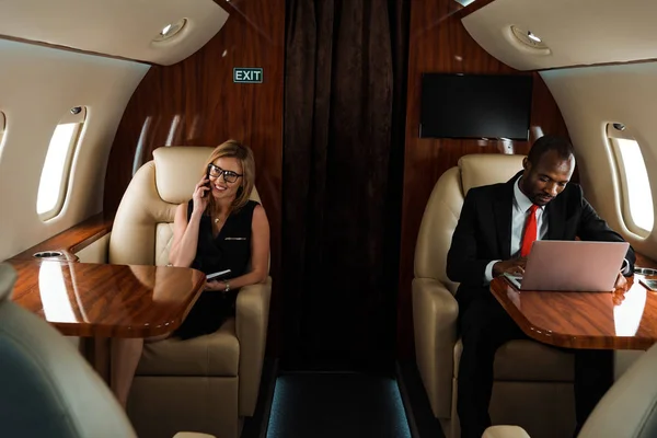 African american businessman using laptop near businesswoman talking on smartphone in private jet — Stock Photo