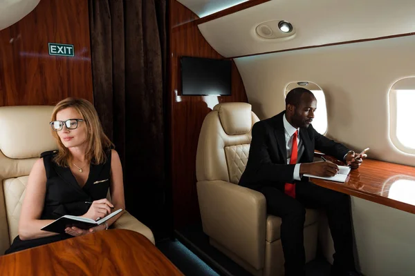 African american businessman using smartphone near businesswoman writing in notebook in private jet — Stock Photo
