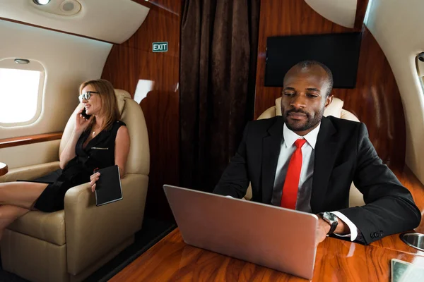 Hombre de negocios afroamericano utilizando portátil cerca de la mujer de negocios hablando en el teléfono inteligente en avión - foto de stock