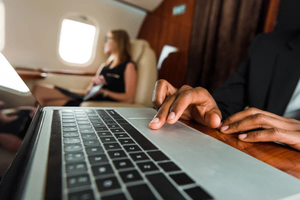 Foco seletivo do empresário afro-americano digitando no laptop perto de empresária no avião — Fotografia de Stock