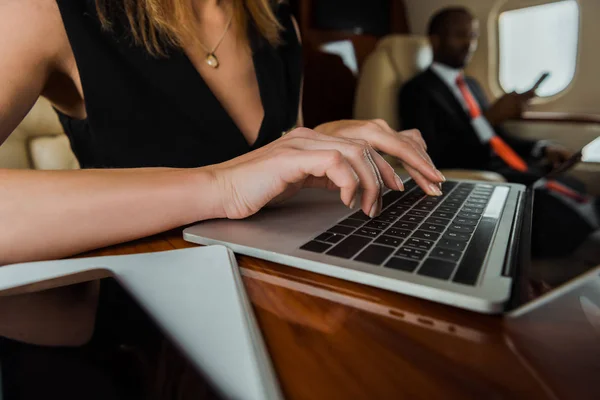 Selective focus of businesswoman typing on laptop near african american businessman in plane — Stock Photo