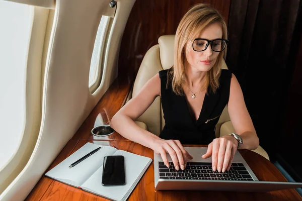 Attractive businesswoman in glasses typing on laptop near smartphone with blank screen — Stock Photo