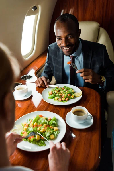 Foco seletivo do empresário afro-americano perto de deliciosos pratos e empresárias em avião privado — Fotografia de Stock