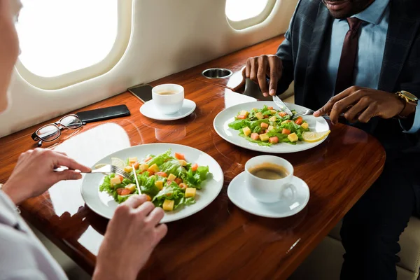 Vista recortada de hombre de negocios afroamericano cerca de sabrosos platos y mujer de negocios en avión privado - foto de stock