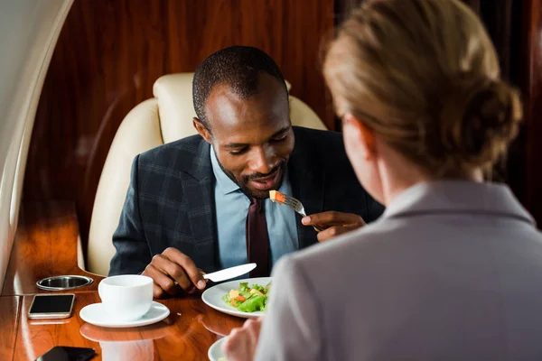 Enfoque selectivo de hombre de negocios afroamericano comiendo cerca de la mujer de negocios y sabrosa ensalada en avión privado - foto de stock