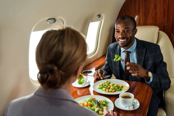 Enfoque selectivo de empresario afroamericano feliz cerca de deliciosos platos y mujer de negocios en avión privado - foto de stock