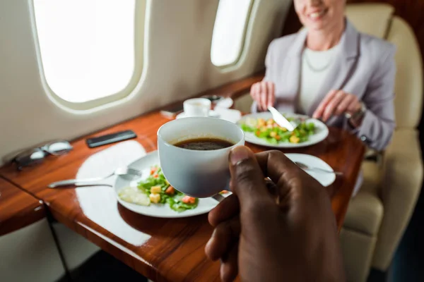 Vista recortada de hombre afroamericano sosteniendo taza con café cerca de la mujer en avión privado - foto de stock