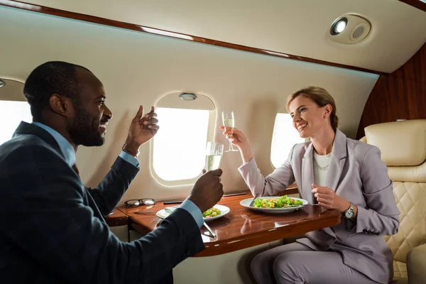 Femme d'affaires heureuse et homme d'affaires afro-américain avec verres à champagne près des salades en jet privé — Photo de stock
