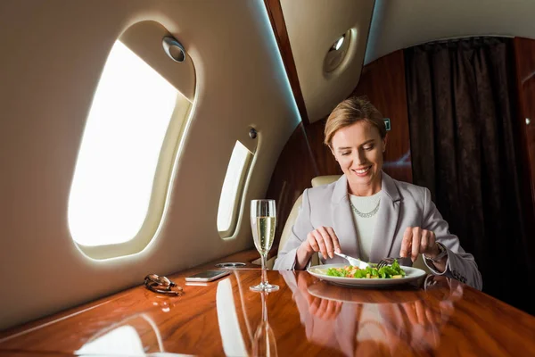 Felice donna d'affari che tiene posate vicino insalata e bicchiere di champagne in jet privato — Foto stock