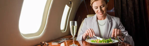 Colpo panoramico di felice donna d'affari in possesso di posate vicino insalata e bicchiere di champagne in jet privato — Foto stock