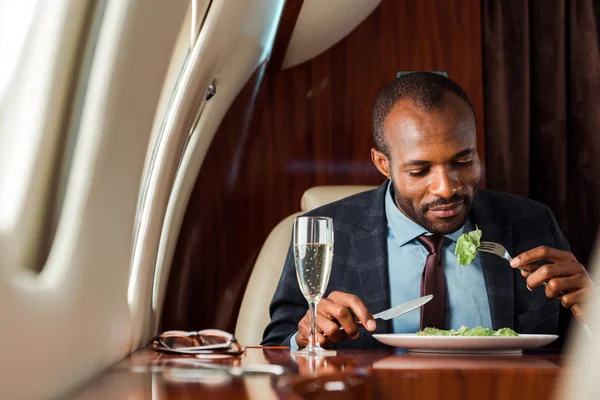 Guapo afroamericano hombre de negocios comiendo ensalada en jet privado - foto de stock