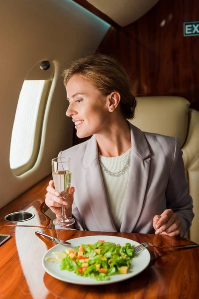 Happy businesswoman with champagne glass near tasty salad in private jet — Stock Photo