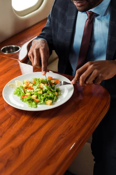 Vista recortada del hombre de negocios afroamericano sosteniendo cubiertos cerca de ensalada en jet privado - foto de stock