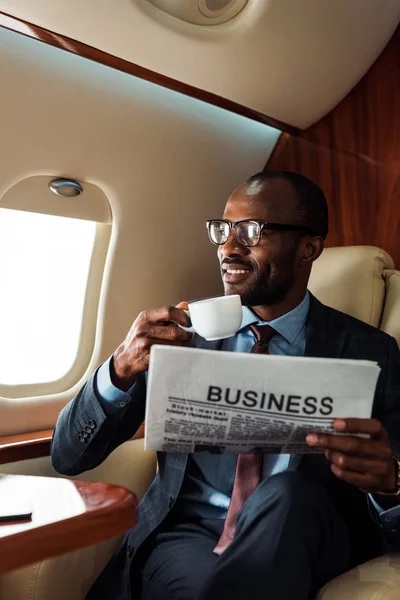 Feliz afroamericano hombre de negocios en gafas celebración de periódico de negocios y taza en avión privado - foto de stock