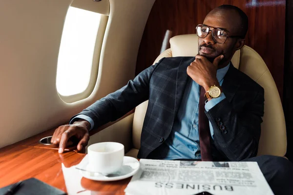 Selektiver Fokus des afrikanisch-amerikanischen Geschäftsmannes in Brille in der Nähe von Wirtschaftszeitung und Tasse im Privatflugzeug — Stockfoto