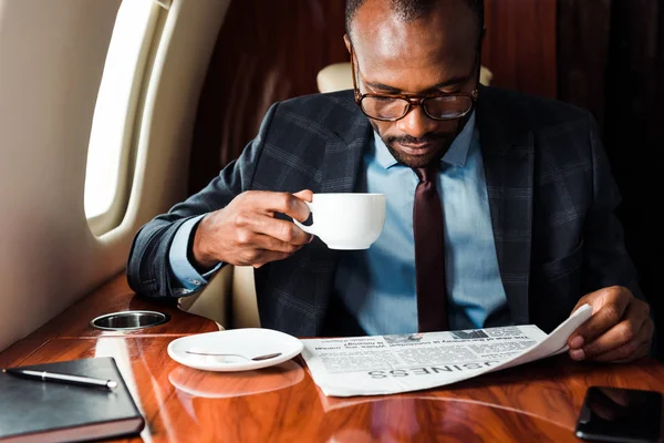 Homme d'affaires afro-américain à lunettes lisant un journal d'affaires tout en tenant une tasse dans un avion privé — Photo de stock
