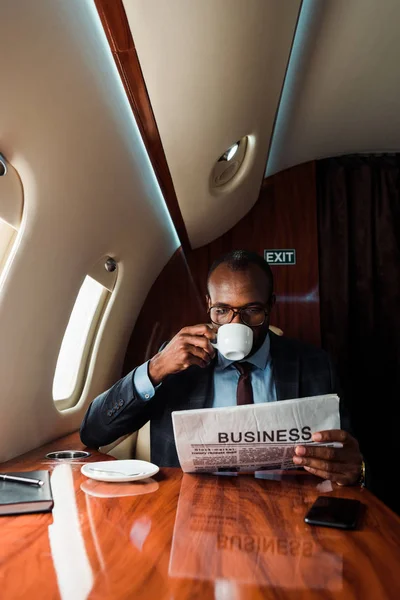 Empresario afroamericano en gafas leyendo periódico de negocios mientras bebe café en avión privado - foto de stock