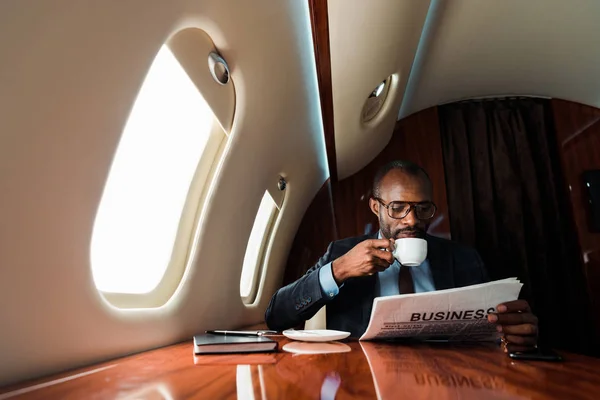 Empresario afroamericano leyendo periódico de negocios mientras bebe café en avión privado - foto de stock