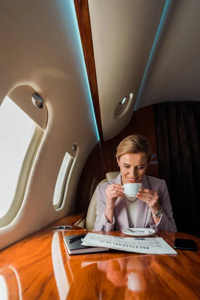 Happy businesswoman holding cup while reading business newspaper in private plane — Stock Photo