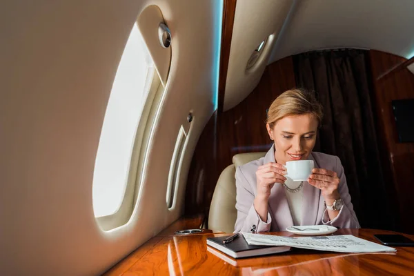 Alegre mujer de negocios sosteniendo taza mientras lee periódico de negocios en avión privado - foto de stock