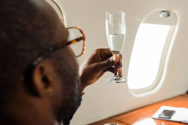 Selective focus of african american businessman in glasses holding champagne in private jet — Stock Photo