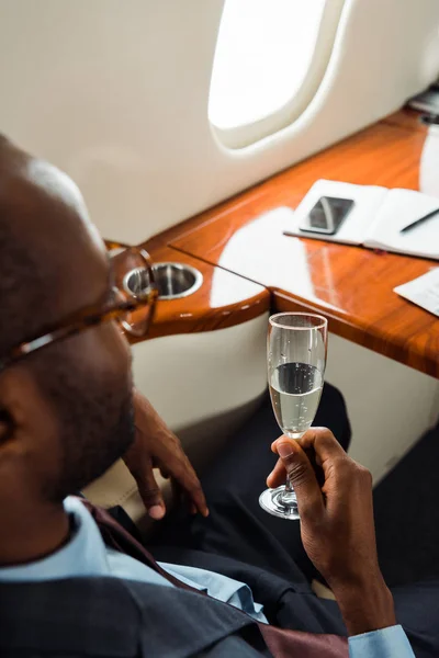 Selective focus of african american businessman in glasses holding champagne glass in private jet — Stock Photo