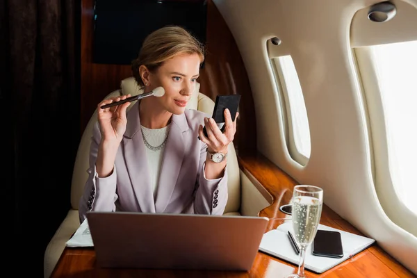 Empresária aplicando cosméticos decorativos perto de gadgets e taça de champanhe em avião privado — Fotografia de Stock