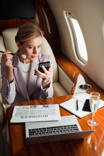 Mujer que aplica cosméticos decorativos cerca de gadgets y copa de champán en avión privado - foto de stock
