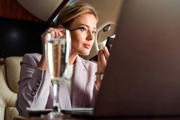 Enfoque selectivo de la mujer de negocios que aplica rímel cerca de la computadora portátil y copa de champán en avión privado - foto de stock