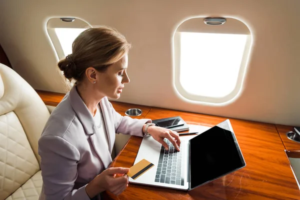 Attractive businesswoman using laptop with blank screen and holding credit card in private plane — Stock Photo