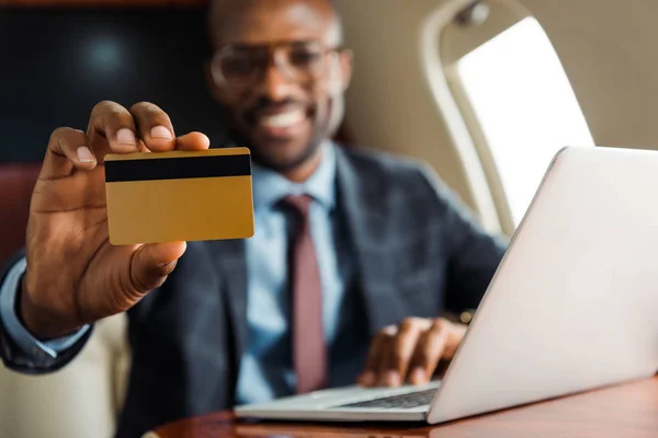 Selective focus of african american businessman holding credit card near laptop in private plane — Stock Photo