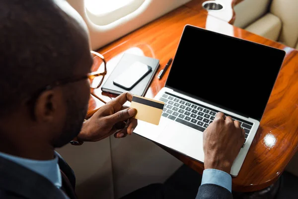 Messa a fuoco selettiva dell'uomo d'affari africano americano che tiene la carta di credito vicino al computer portatile con schermo vuoto in aereo privato — Foto stock