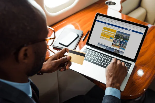 Selective focus of african american businessman holding credit card near laptop with booking website in private plane — Stock Photo