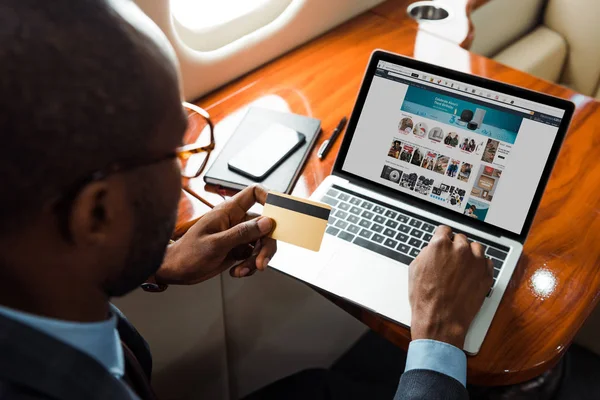 Selective focus of african american businessman holding credit card near laptop with amazon website in private plane — Stock Photo