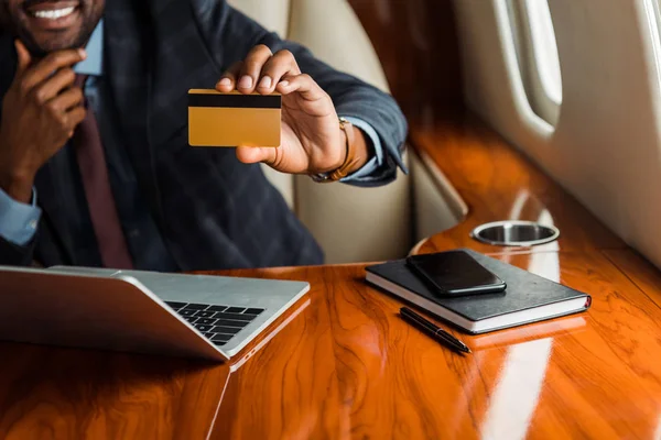 Vue recadrée d'un homme d'affaires afro-américain tenant une carte de crédit près d'un ordinateur portable et d'un smartphone dans un avion — Photo de stock
