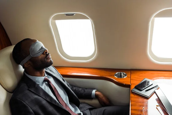 Африканский американский бизнесмен в сонной маске сидит рядом с гаджетами в частном самолете — стоковое фото