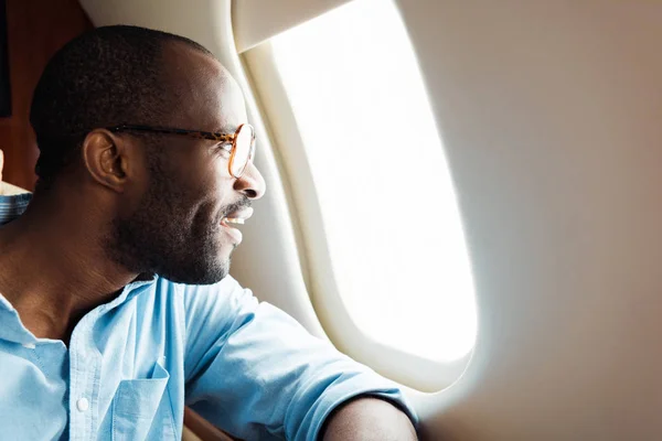 Homem americano africano feliz em óculos olhando para a janela do avião — Fotografia de Stock